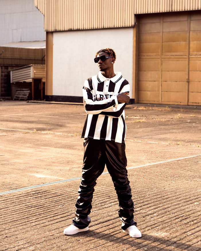about-05 Fashionable man in black and white attire posing confidently outdoors in an industrial area.