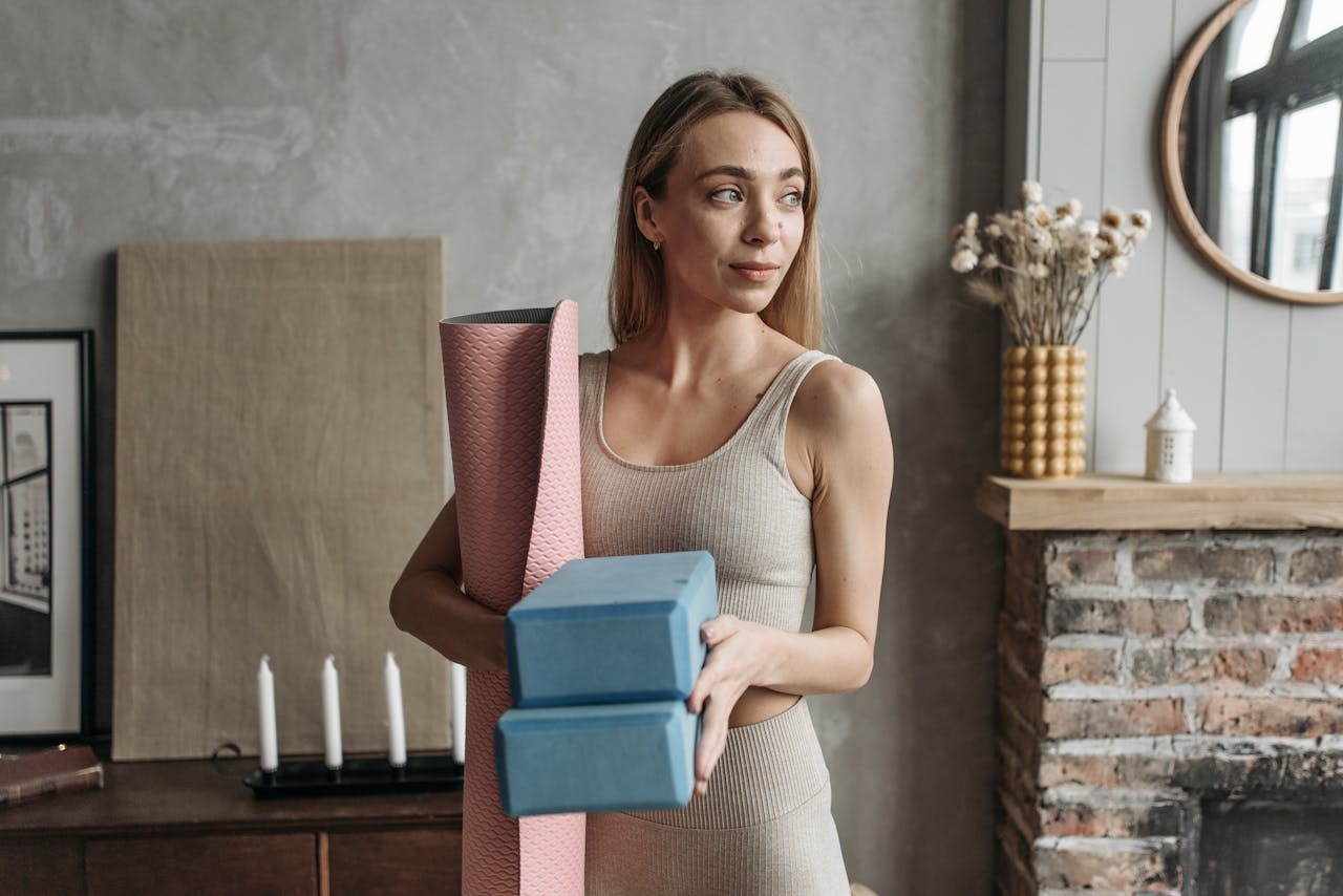 services-06 A woman in cozy activewear holding yoga blocks and mat indoors, ready to exercise.